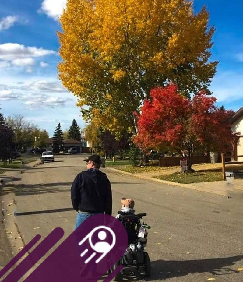 A person walks beside a child in a wheelchair down a quiet residential street lined with vibrant yellow and red autumn trees under a blue sky.